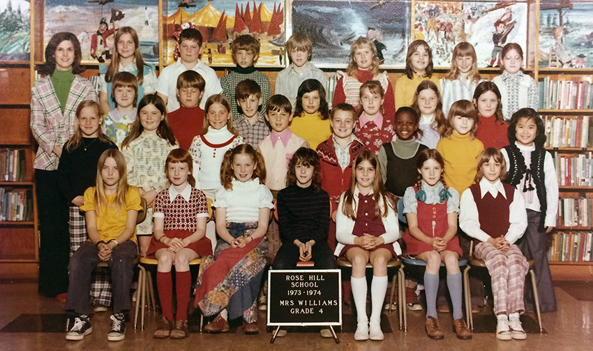 Color class photograph from the 1973 to 1974 school year showing Mrs. Williams' fourth grade class. There are 30 students pictured, mostly girls. Nearly all of the children are Caucasian except for one African-American child and one Asian child. Mrs. Williams is standing in the back left corner. She is wearing a pink, black, and white striped jacket over a green turtleneck shirt. 