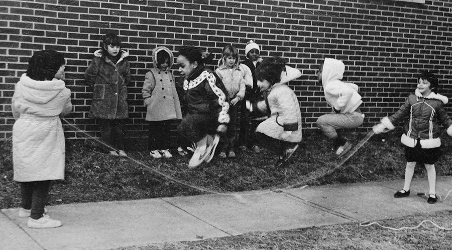 Black and white photograph from the cover of Rose Hill Elementary School's 1982 to 1983 yearbook showing a group of students playing jump rope on the sidewalk. Ten girls are pictured, five of whom are waiting along the wall for their turn. Three girls are in mid-air as two other girls swing the rope beneath them. 