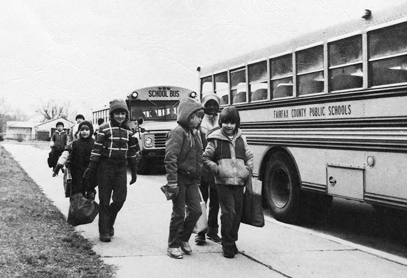 Black and white photograph from the cover of Rose Hill Elementary School's 1983 to 1984 yearbook showing a group of students walking up the sidewalk toward the entrance of the school after having been dropped off by their school buses. Two buses are visible on the right. 