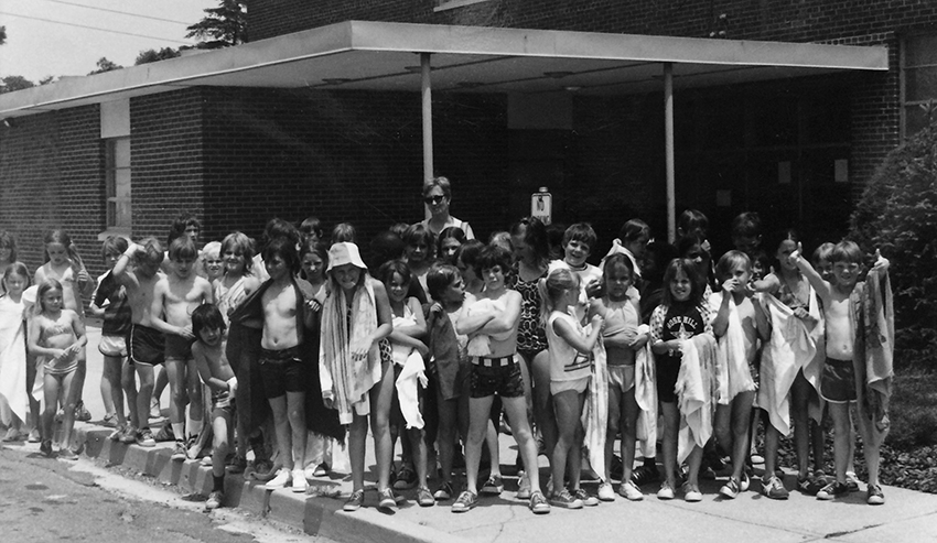 Black and white photograph of a group of about 41 students, boys and girls, standing in front of the entrance to Rose Hill Elementary School. The children are wearing bathing suits and holding towels. A female teacher is standing at the back of the group. 